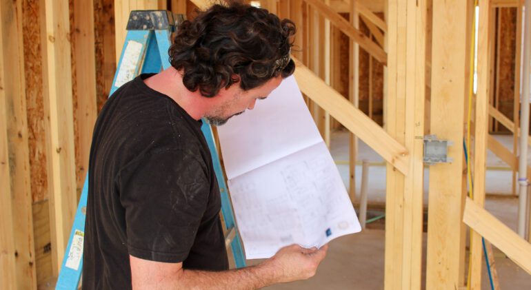 "Construction worker reviewing blueprint inside framed home, planning next steps for building project."