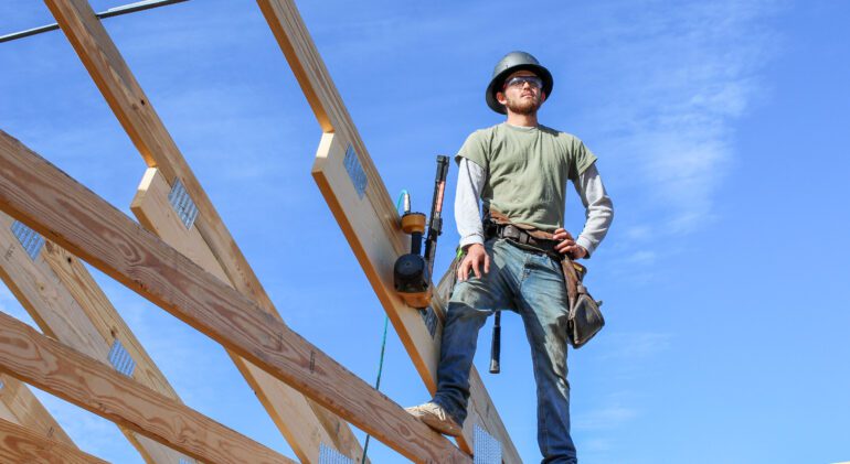 "Roofer standing on home frame wearing tool belt, preparing to build roof, construction site in progress."