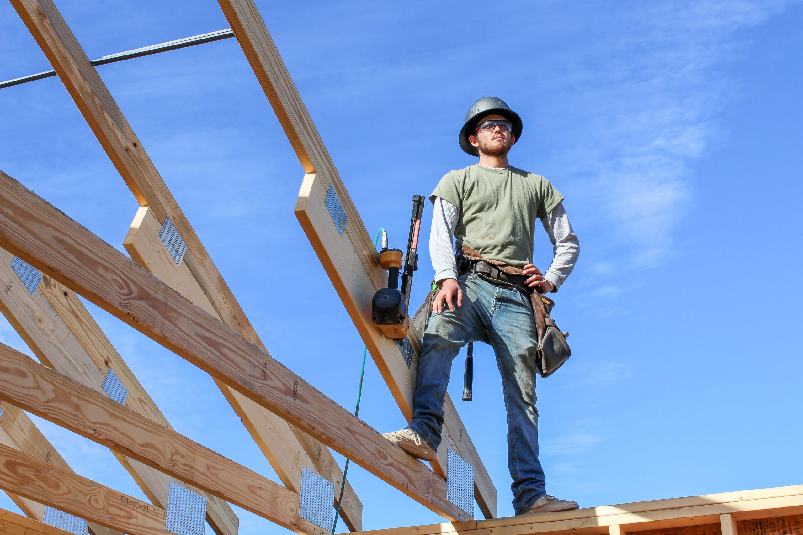 "Roofer standing on home frame wearing tool belt, preparing to build roof, construction site in progress."